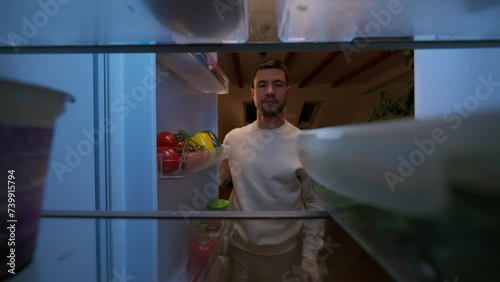View inside kitchen fridge: handsome man opens fridge door, putting inside salad in box