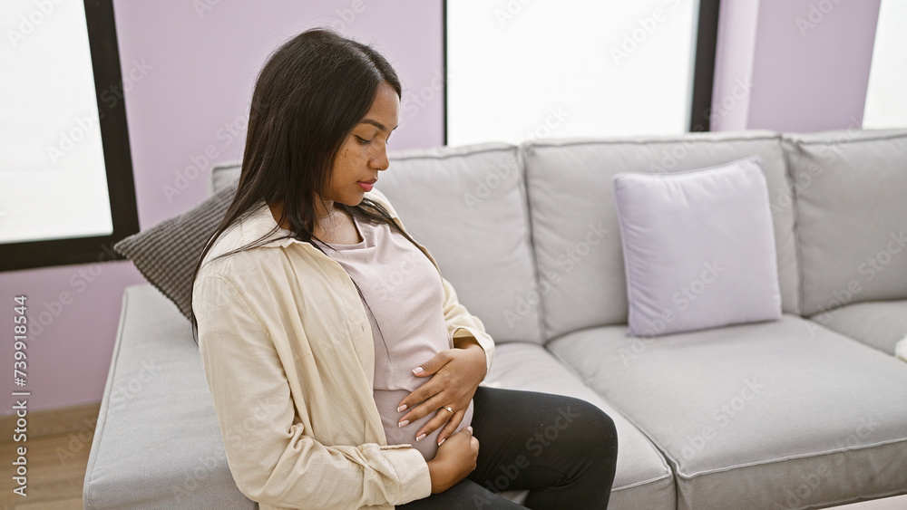 Serious-faced, pregnant young woman sitting at home, casually touching her belly on her living room sofa, embodying the emotion and relaxed anticipation of motherhood. Serious-faced, pregnant young woman sitting at home, casually touching her belly on her living room sofa, embodying the emotion and relaxed anticipation of motherhood.