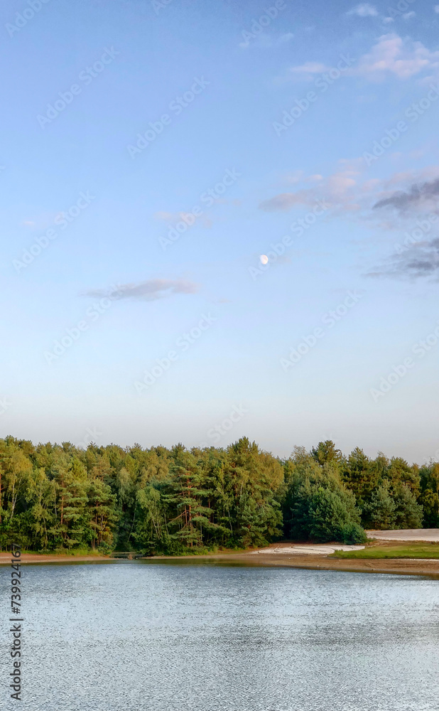 The image showcases a tranquil lake with a gentle ripple on the water surface, reflecting the waning light of dusk. A dense forest of coniferous trees forms a dark green band that separates the lake The image showcases a tranquil lake with a gentle ripple on the water surface, reflecting the waning light of dusk. A dense forest of coniferous trees forms a dark green band that separates the lake