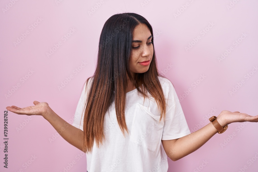 Young arab woman standing over pink background smiling showing both hands open palms, presenting and advertising comparison and balance Young arab woman standing over pink background smiling showing both hands open palms, presenting and advertising comparison and balance