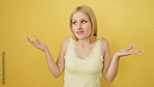 A perplexed young blonde woman shrugging against a yellow background, epitomizing confusion.