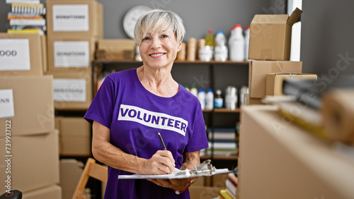 Mature woman volunteering at a warehouse takes notes among labeled boxes indoors.