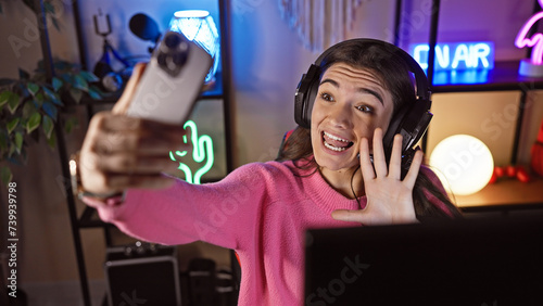 Young hispanic woman streaming videogames in a dark room, wearing headphones, waving at webcam, neon lights background.