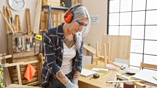 Serious middle age grey-haired woman carpenter working industriously, donned in safety headphones and security glasses at her indoor carpentry workshop.
