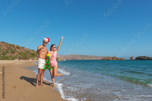 Happy couple in Santa hat walking on the beach with inflatable Christmas tree. Christmas vacation concept.