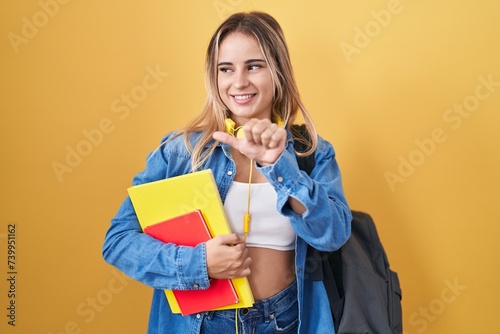 Young blonde woman wearing student backpack and holding books pointing to the back behind with hand and thumbs up, smiling confident