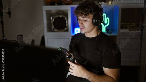 In the heat of the night, handsome, young hispanic streamer deep in virtual gaming, headphones on, fielding video call amidst serious gaming room action photo