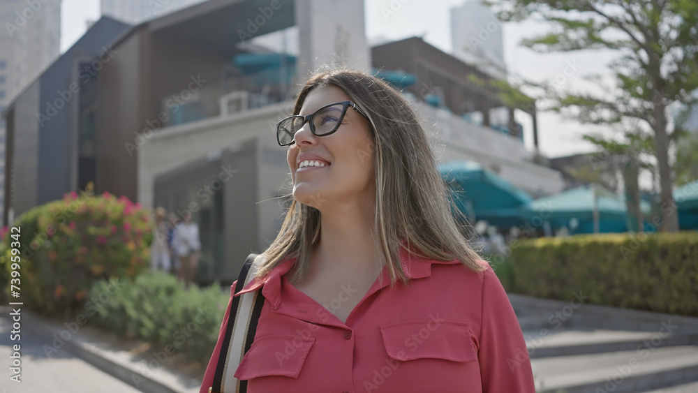 A smiling young adult hispanic woman with brunette hair wearing glasses in a dubai city marina setting.