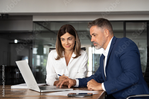 Team of diverse partners sitting at table mature Latin business man and European business woman discussing project on laptop in office. Two colleagues of professional business people working, close up
