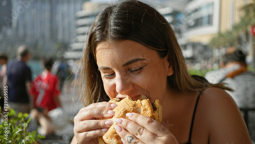 Fotografia A young hispanic woman enjoying a sandwich outdoors with skyscrapers, including the burj khalifa, in dubai
