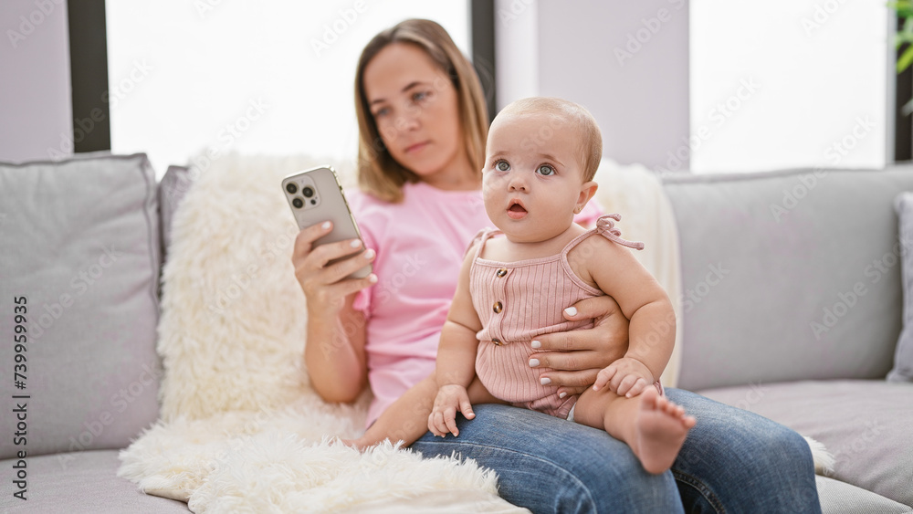 Relaxed mother, sitting on the sofa, having a serious expression as she texts on her smartphone, sharing a lovely moment with her infant daughter at home Relaxed mother, sitting on the sofa, having a serious expression as she texts on her smartphone, sharing a lovely moment with her infant daughter at home