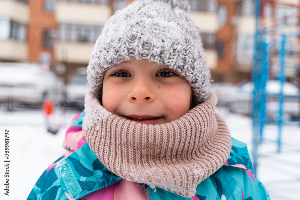 Smiling child in a knitted hat and scarf outdoors with a snowy playground in the background Smiling child in a knitted hat and scarf outdoors with a snowy playground in the background
