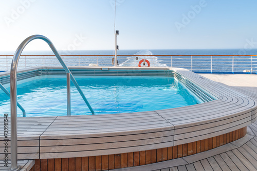 A swimming pool with clear, clean water at the stern of a cruise ship with a view of the ship's wake stretching to the horizon. Wooden deck, handrails and lifebuoy.