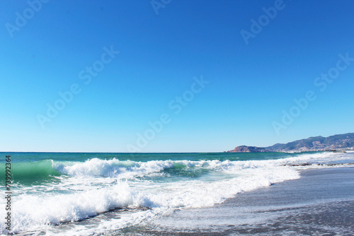 Fototapeta Naklejka Na Ścianę i Meble -  clear blue sky and waves with white foam at Mahmutlar beach in Turkey. mountains in the background