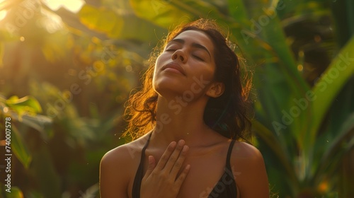 Young latin woman with hand on chest breathing in fresh air in a beautiful garden during sunset. Healthy mexican girl enjoying nature while meditating during morning exercise routine with closed eyes photo