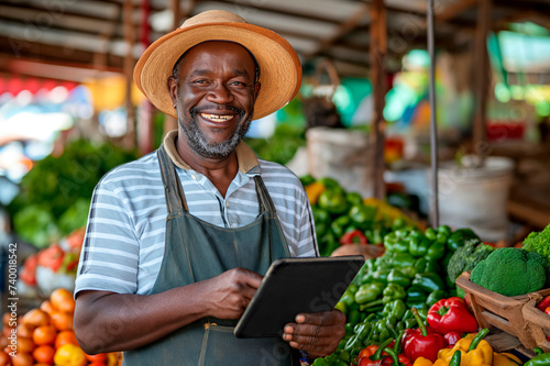 portrait of a smiling african farmer at the market, standing with a digital tablet studying the availability of vegetables in the shop