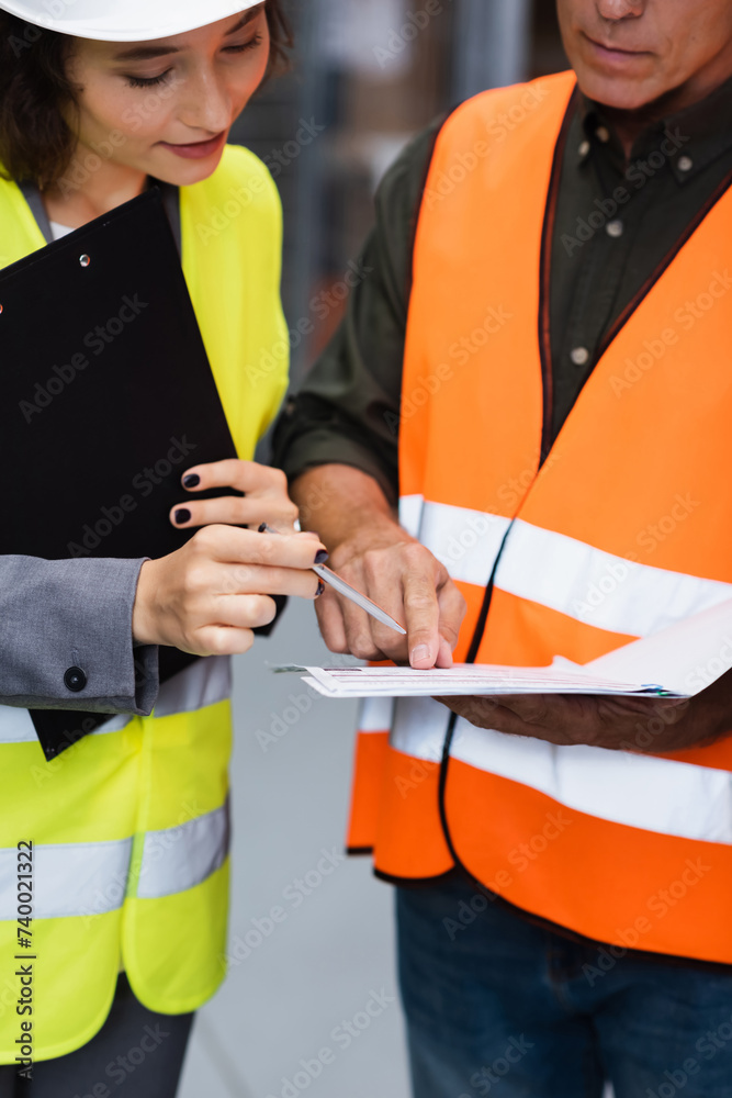 middle aged warehouse supervisor in safety vest showing paperwork to his female employee with pen