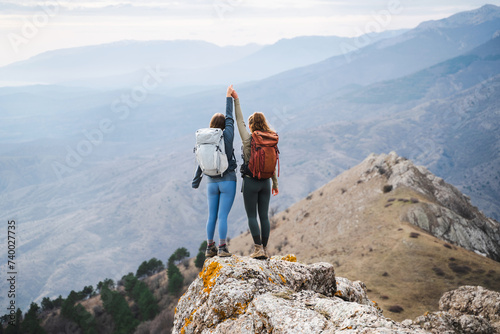 Back view of two young women hikers with arms up with backpacks standing on cliff's edge among the high mountains and looking to the sky on a sunny day, friendship travel tourism adventure concept