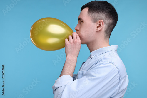 Young man inflating golden balloon on light blue background