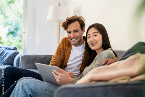 Adult couple looking at the camera while sitting on sofa using digital tablet