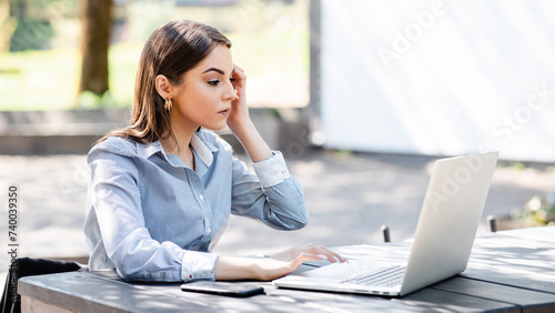 European businesswoman typing on laptop computer outdoors. Beautiful young girl sit at table in sunny park. Modern woman lifestyle. Concept of remote and freelance work