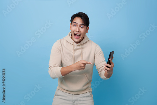Wow face of Asian man shocked what he see and pointing finger in the smartphone isolated on blue background.