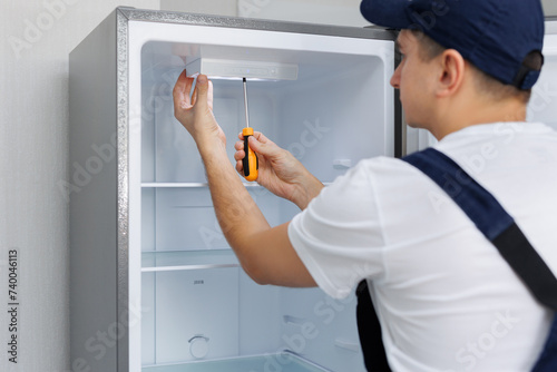 Man in a uniform repairs the light in the refrigerator with a screwdriver. Replacing the light bulb. photo