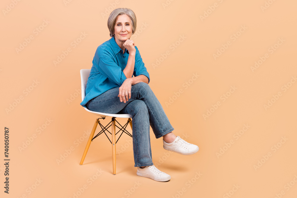 Full length photo of clever thoughtful person wear blue shirt jeans sitting on chair on interview isolated on beige color background Full length photo of clever thoughtful person wear blue shirt jeans sitting on chair on interview isolated on beige color background