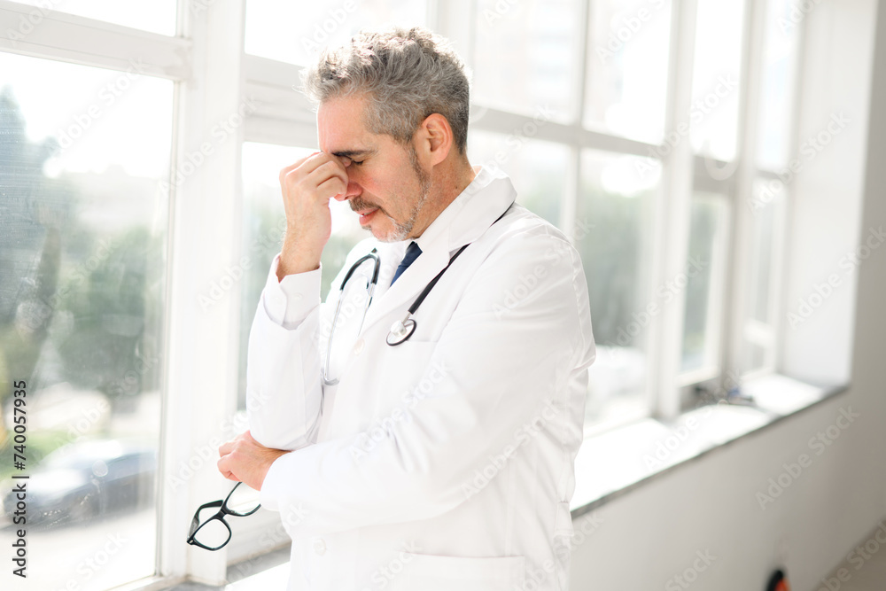 A senior doctor appears in deep contemplation, touching his temple, against the blurred background of a clinic. Pensive face highlights the thoughtful nature of medical decision-making