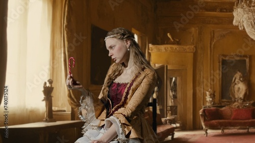Woman in ancient outfit on background of historic interior. Female in renaissance style dress sitting holding candy cane eating sweets enjoying.