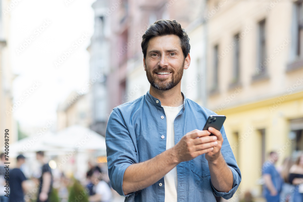 Portrait of a smiling young man standing on a city street, looking at the camera and holding a mobile phone Portrait of a smiling young man standing on a city street, looking at the camera and holding a mobile phone