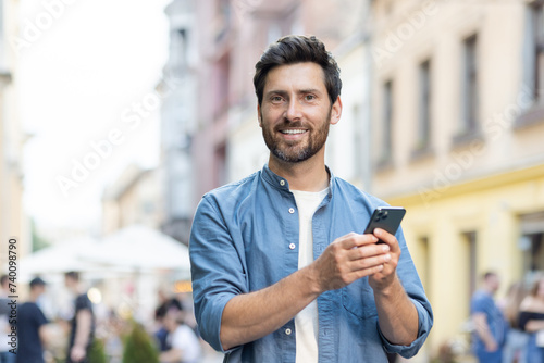 Portrait of a smiling young man standing on a city street, looking at the camera and holding a mobile phone