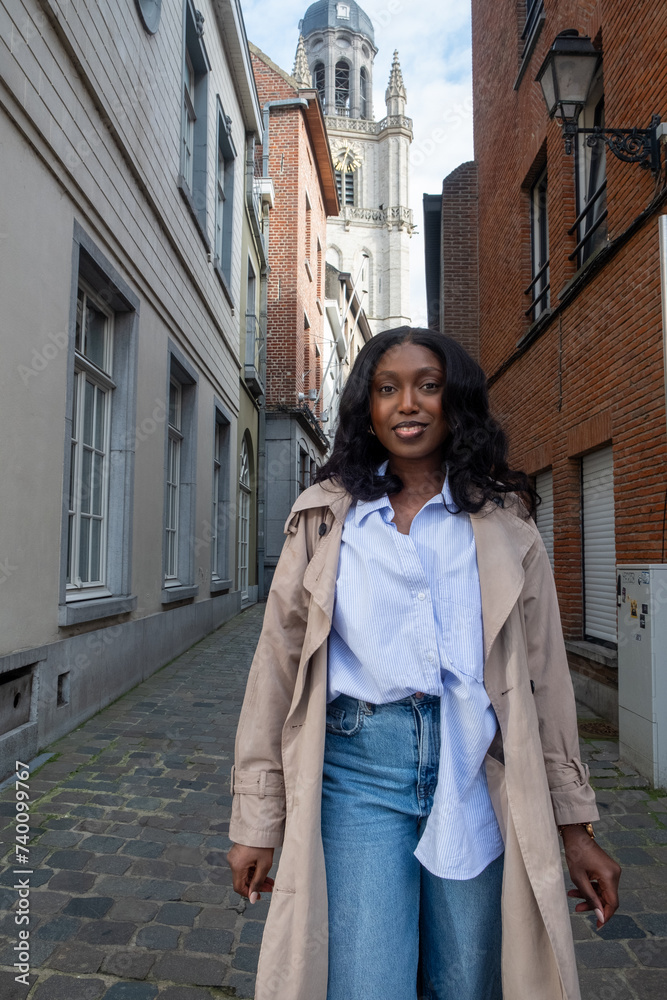 This image depicts a confident young African American woman walking down a cobblestone street in an urban European setting. A historical church tower looms in the background, juxtaposed with the