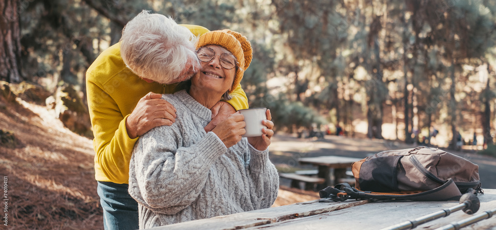 Head shot portrait close up of one middle age woman drinking coffee or tea from a cup sitting at table in the nature in the forest of mountain with old husband hugging wife from the back.