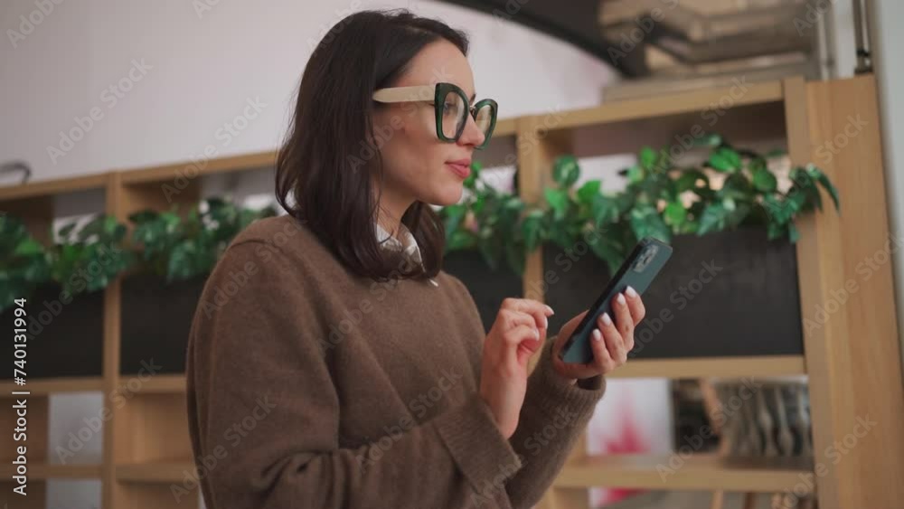 Confident businesswoman in smart glasses talking on smartphone in office. Caucasian female executive text messaging via mobile phone near green plants. Young office worker in sweater. Side view. Confident businesswoman in smart glasses talking on smartphone in office. Caucasian female executive text messaging via mobile phone near green plants. Young office worker in sweater. Side view.