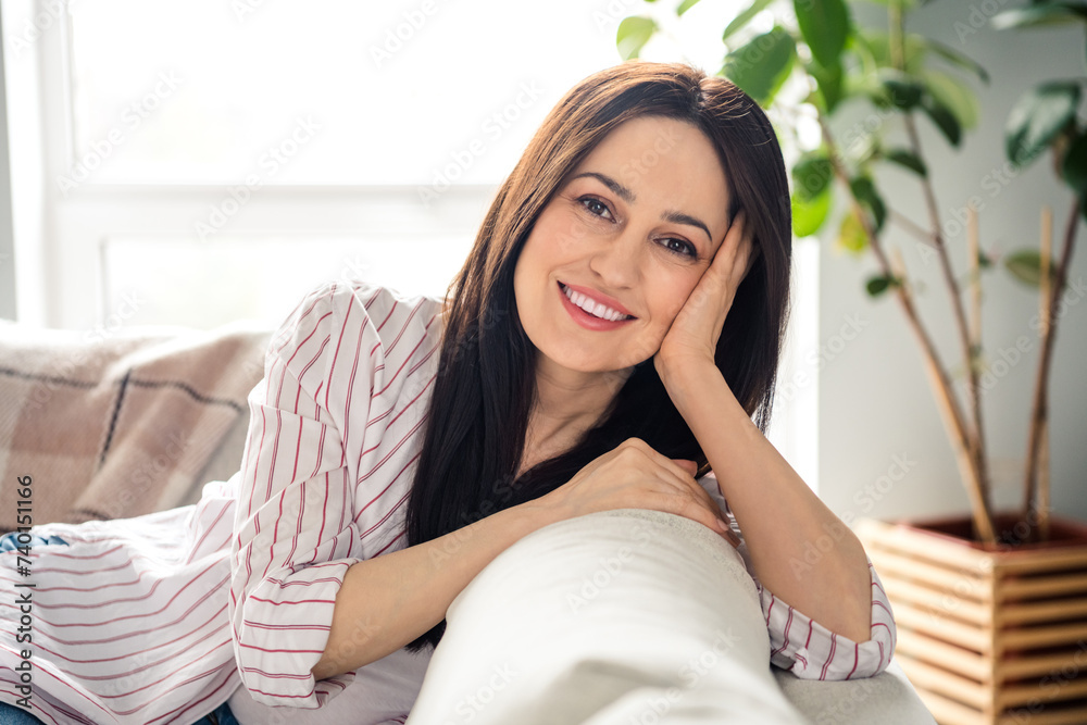 Photo of pretty cheerful carefree girl beaming smile sit couch hand touch cheek sunlight window apartment inside