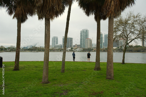 Two men framed by palm trees fishing with poles looking towards downtown. View looking southwest over seawall at Vinoy Park Marina in St. Petersburg, Florida. With palm trees on right and left side. photo