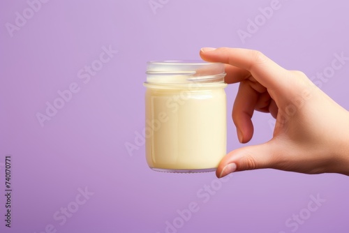 Close-up photo of a devious hand reaching for a jar of mayonnaise cleverly disguised as vanilla pudding for an April Fools' Day gag, against a solid pastel lavender backdrop