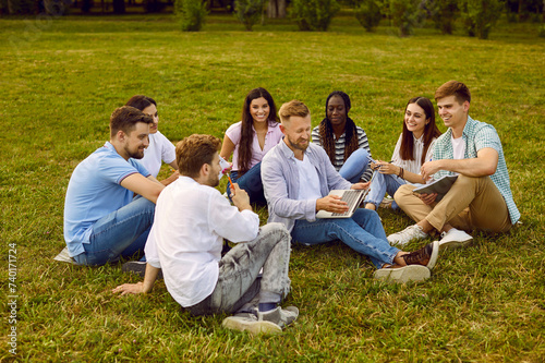 Diverse team of business students and friends working on project in nature. Group of happy young multiracial people sitting on green grass in summer park and using laptop computer together