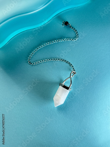 White crystal pendant, pendulum on a chain, healing amulet, spiritual energy detector on wavy blue paper background.
