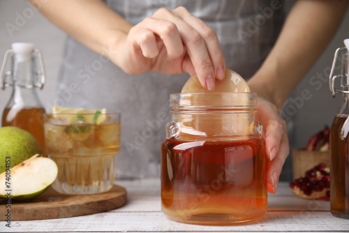 Woman putting Scoby fungus into jar with kombucha at white wooden table, closeup