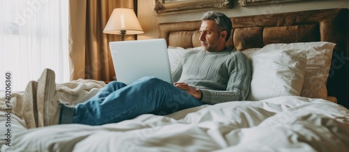 A middle-aged man wearing a sweater and blue jeans sits on a hotel bed, using a laptop to engage in activities related to business, travel, tourism, loneliness, and stay-at-home orders.