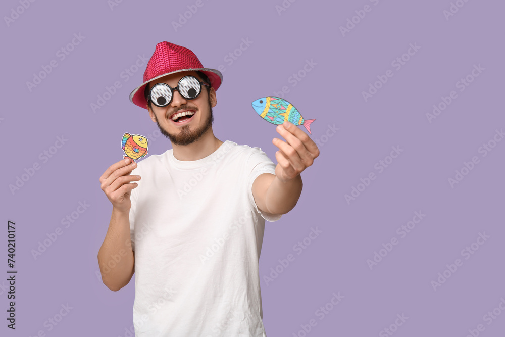 Handsome young man in funny disguise with paper fishes on purple background. April fool's day celebration