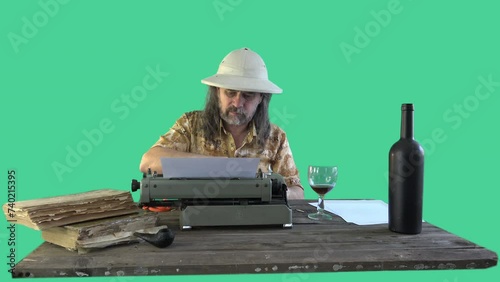 Portrait male traveler (in pith helmet) with long hair writes his memoirs at an old typewriter, contentedly drinks wine from glass at wooden table, there are books and blank sheets of paper on table photo