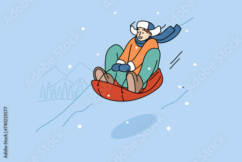 Teenage boy is relaxing at winter ski resort, sliding down snow-covered hill on inflatable sled. Happy young guy leads active lifestyle in winter season, during vacation or holidays
