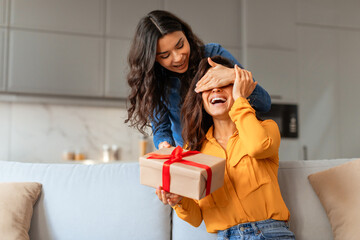 lady surprises girlfriend playfully covering eyes before giving gift indoor