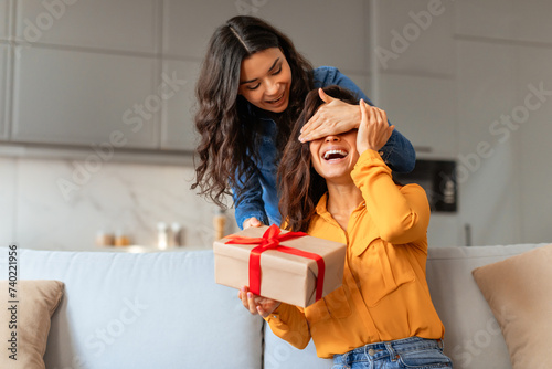 lady surprises girlfriend playfully covering eyes before giving gift indoor