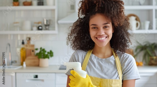 A happy young woman smiles while standing in a white kitchen, wearing yellow cleaning gloves and holding a cleaning agent