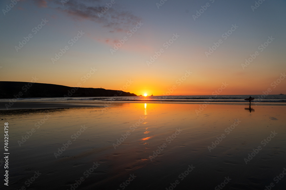 Silhouette d'un surfeur marchant sur le sable mouillé, capturé au coucher du soleil sur la plage des Trépassés, dans le Finistère en Bretagne : une scène empreinte de magie et de liberté.