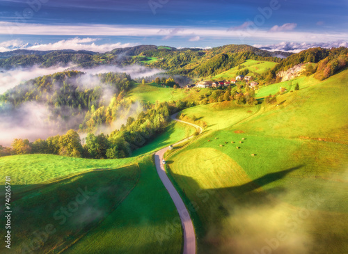 Aerial view of country road in green alpine meadows at sunrise in summer. Top drone view of rural road, mountains in low clouds. Colorful landscape with village, hills, fields, green grass. Slovenia